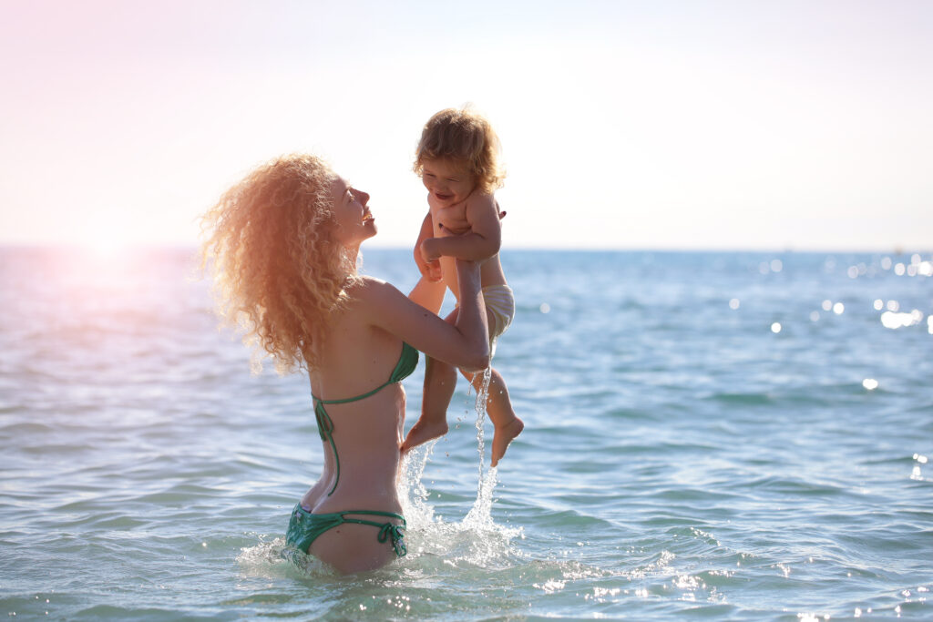 Mom carrying baby in the ocean wearing bikini showing off her mommy makeover results