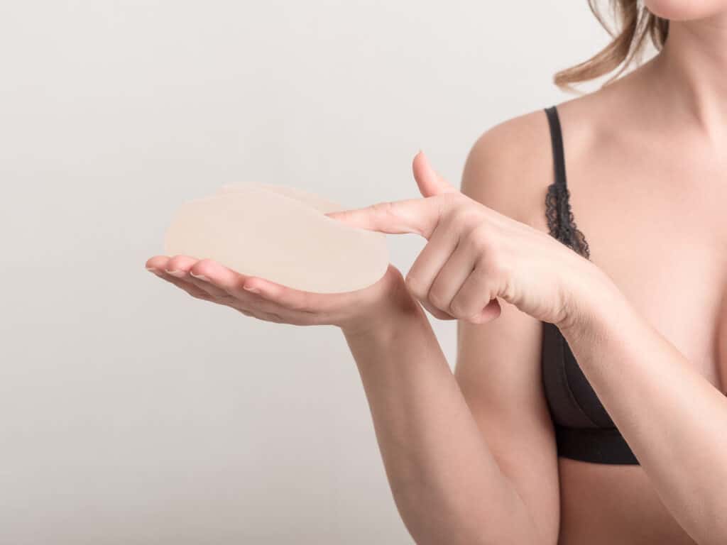 Woman, wearing black bra, holds a breast implant in her hand during her explant surgery consultation